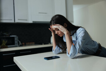 Stressed woman sitting on kitchen counter with phone in front of her, feeling overwhelmed and exasperated as she holds her head in hands - Powered by Adobe