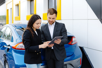 Smiling woman standing at the car and pointing at car seller tablet. She is picked right car for her she saw online. A woman concludes an agreement to buy a car and shakes the manager's hand