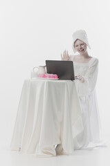 Young attractive woman dressed as simple peasant woman waving hello while have meeting online at laptop against white studio background. Concept of renaissance art, comparison of eras, vintage. Ad