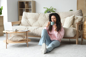 Young African-American woman with cup of coffee resting on floor at home