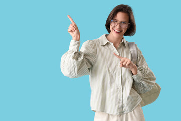 Happy female student with backpack pointing at something and winking on blue background