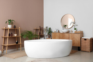 Interior of stylish bathroom with bathtub, houseplants and wooden furniture