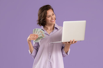 Pretty young woman with money and laptop on lilac background