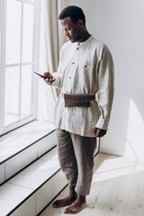 Stylishly dressed African American man stands by large window, absorbed in using his smartphone, in spacious and well-lit modern home setting. Clothes made of natural linen fabric, eco-friendly shirt.