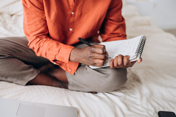 Focused African American man sits on bed, jotting down notes in a notebook, with laptop nearby, in comfortable home setting .