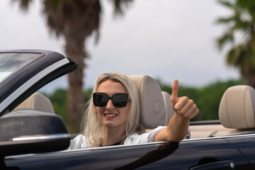 Portrait of a young smiling blonde woman in sunglasses driving a convertible convertible showing a...