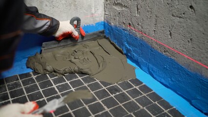 A worker applies adhesive to the floor with a scraper before gluing the tiles. A close-up of a...
