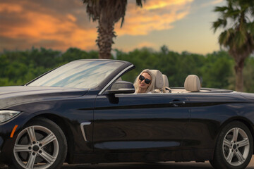 Smiling young beautiful blonde woman in sunglasses is sitting at the wheel of a dark blue...