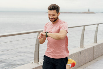 A smiling man checks his smartwatch during a seaside jog, dressed in a pink shirt and running...