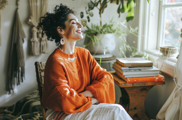 A woman in an orange sweater and white pants sits on her chair, smiling as she meditates with...