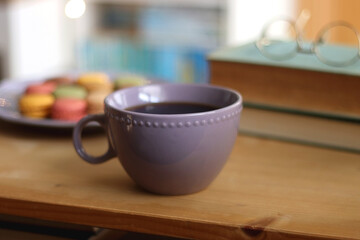 Purple plate filled with pastel macarons, cup of tea or coffee, vintage books and reading glasses on the table. Colorful bookcase in the background. Selective focus.