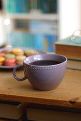 Purple plate filled with pastel macarons, cup of tea or coffee, vintage books and reading glasses on the table. Colorful bookcase in the background. Selective focus.
