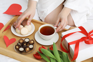 Tasty breakfast served in bed. Woman with tea, desserts, gift box and flowers at home, closeup