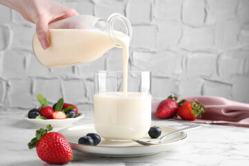 Woman pouring tasty yogurt into glass at white marble table, closeup
