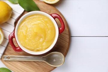 Delicious lemon curd in bowl, fresh citrus fruits and spoon on white wooden table, flat lay. Space...