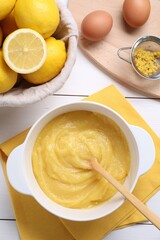 Delicious lemon curd in bowl, ingredients, spoon and sieve on white wooden table, flat lay