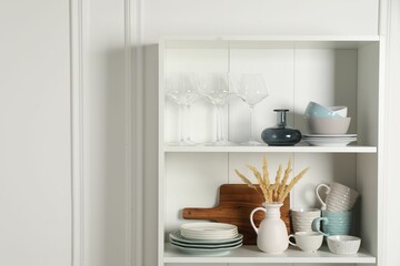 Different ceramic dishware and glasses on shelves in cabinet indoors. Space for text