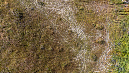 Aerial view of an abandoned soccer field. A lone goalpost stands amidst overgrown grass. Circular motorbike tracks crisscross the field, indicating where nature and human activity intersect.