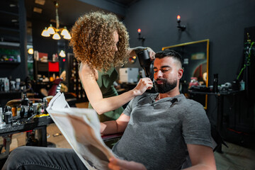 In a stylish barbershop, a barber skillfully dries and styles a male client's hair as he comfortably reads, blending relaxation with grooming elegance.