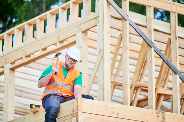 Carpenter constructing two-story wooden frame house. Bearded man in glasses hammering nails into...