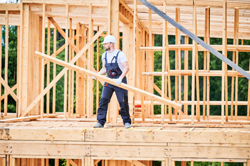 Carpenter constructs wooden-framed house. Bearded man holds large truss in his hands while dressed in work clothes and helmet. An idea of modern and eco-friendly construction.
