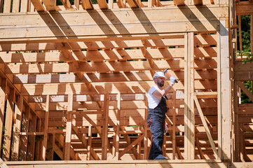 Carpenter constructing wooden frame, two-story house. Bearded man hammering nails with hammer,...