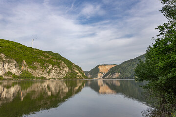 Obraz premium natural landscape featuring a calm lake with reflections of the surrounding greenery and cliff. A wind turbine is perched atop the cliff, indicating a commitment to renewable energy.