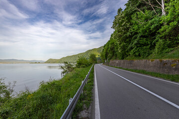  A winding asphalt road runs alongside a serene lake, bordered by a guardrail and lush greenery....