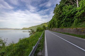 Obraz premium A winding asphalt road runs alongside a serene lake, bordered by a guardrail and lush greenery. The tranquil scene is framed by a mountain range under a blue sky dotted with white clouds.