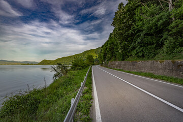 Obraz premium A winding asphalt road runs alongside a serene lake, bordered by a guardrail and lush greenery. The tranquil scene is framed by a mountain range under a blue sky dotted with white clouds