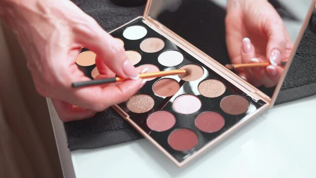 Crop woman hand taking eyeshadow shade from palette in brush. Anonymous female is using colorful make-up on table.