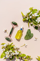Set of natural jade cosmetic massager and gua sha scraper, oil for lymphatic drainage massage on beige vertical background. branches of a flowering.