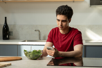 A young man is seated at a kitchen table, engrossed in his cell phone. He appears focused as he...