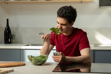 A young man in a red shirt sits at a kitchen counter with a bowl of fresh, green salad, expressing...