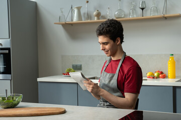 A young man wearing a grey apron over a casual outfit is sitting in a well-lit modern kitchen. His...