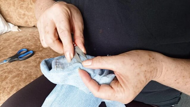 Horizontal view of a woman's hands using sewing scissors to break the threads of a piece of jean fabric to remove a patch. Clothing recycling.