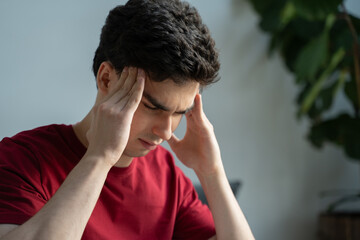 A young man wearing a red shirt is visibly distressed as he grips his temples tightly, expressing...