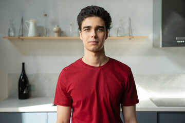 A young man stands confidently in a modern kitchen setting, wearing a casual red T-shirt. The...
