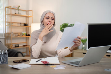 Young worried and shocked Muslim woman in hijab holding documents and calculator. Financial problems with accounts, loans, family budget at the home