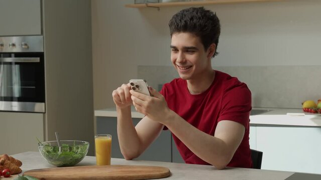 A young man is seated at a kitchen table, engrossed in his cell phone. He appears focused as he scrolls through the screen, unaware of his surroundings.