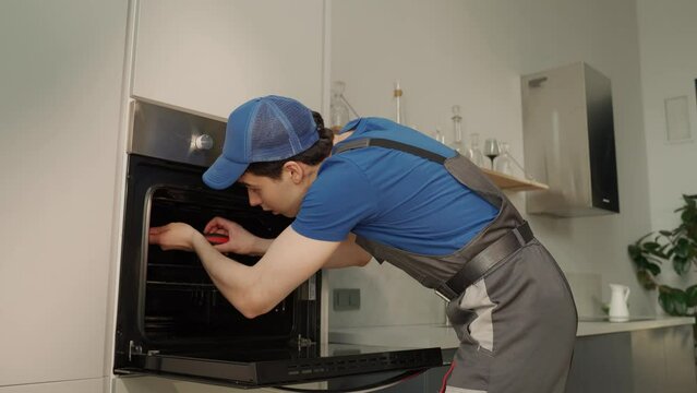 A professional appliance repairman, dressed in a blue shirt and protective workwear, is focused on fixing a contemporary oven built into sleek kitchen cabinetry. With expert care, he adjusts the