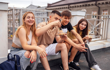 Four teenagers are sitting on the parapet on the roof of the house and eating Asian food. Two young couples snack on fast food and have fun together.