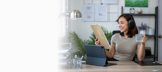 A woman is sitting at a desk with a laptop and a tablet. She is smiling and holding a glass of...