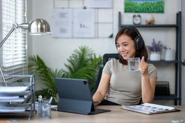 Fototapeta premium A woman is sitting at a desk with a laptop and a glass of water. She is wearing headphones and she is enjoying her work