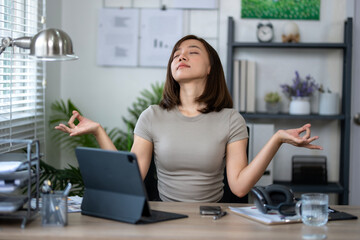 A woman is sitting at a desk with a tablet and a laptop. She is wearing a grey shirt and she is in a relaxed state