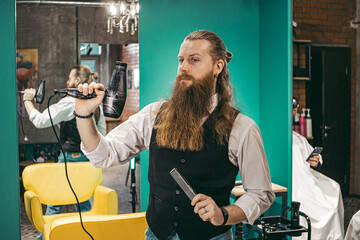Hairdresser funny shoots a hairdryer. Man young barber barber cheerful joking with long beard and...