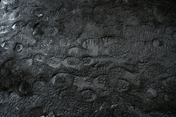 Black sand texture with shoe footprints. Wet volcanic soil, traces of human passage. Reflections of light, water, beach, mud.