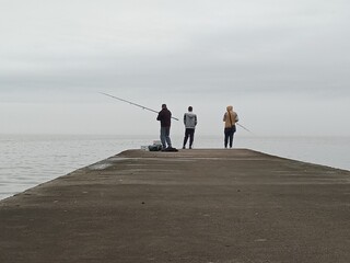 Three friends fishing on a breakwater on a completely cloudy day. Fishermen. Friendship