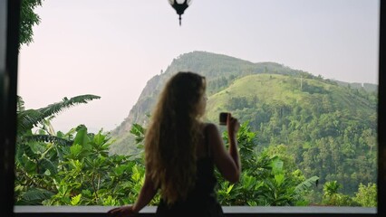 Blonde woman sips coffee at upscale resort balcony with breathtaking Ella Rock mountain landscape in backdrop. Leisurely enjoying morning amidst Sri Lankan green plants, embodies serene travel luxury.