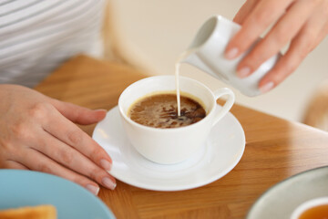 Breakfast. Woman pouring cream into cup of coffee at wooden table, closeup